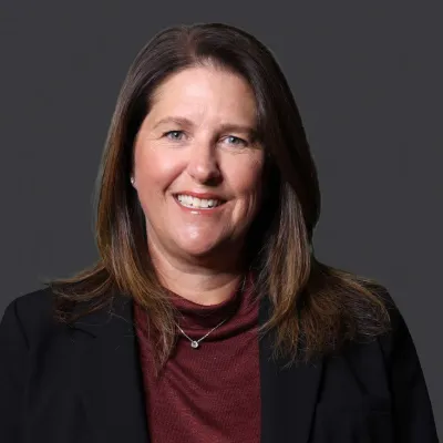 Smiling middle-aged woman with brown hair wearing a black blazer and burgundy top against a dark background