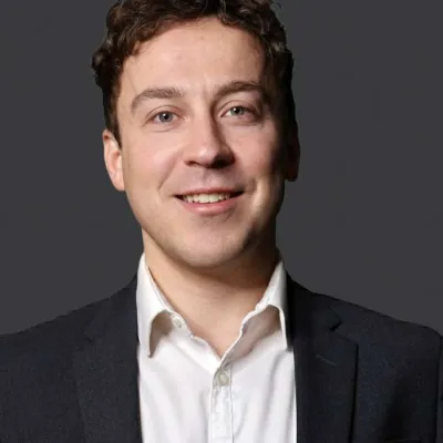 Smiling young man with curly hair in white shirt and dark blazer against gray background