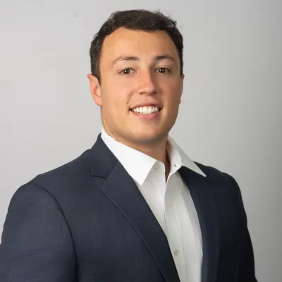 Young professional man in navy suit and white shirt smiling with hands clasped against gray background