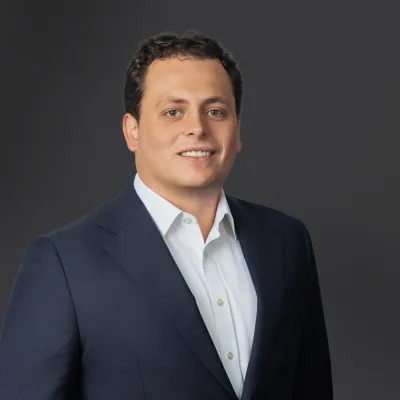 Professional headshot of a man in a navy suit and white shirt smiling against a dark gray background
