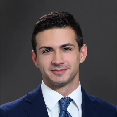 Professional headshot of a young man in a suit with a blue tie on a gray background.