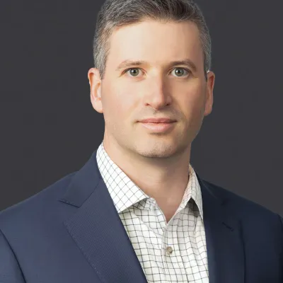 Professional headshot of a man with gray hair wearing a navy blazer and checkered shirt against a gray background