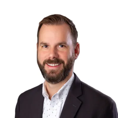 Smiling man with beard wearing a dark blazer and patterned white shirt on white background