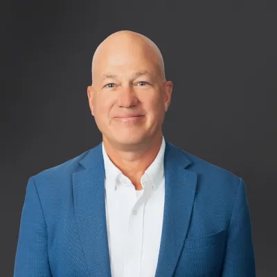 Smiling bald man wearing a blue blazer and white shirt against a dark gray background.