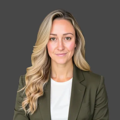 Professional young woman with long blonde hair wearing an olive blazer and white top against a gray background