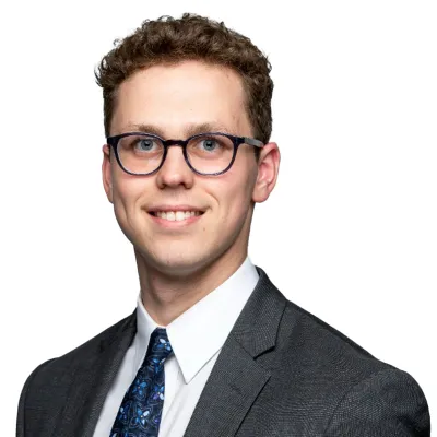 Professional portrait of a young man in a suit with glasses and a tie, smiling confidently at the camera.