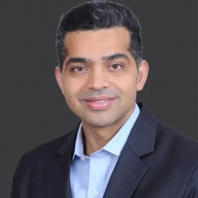 Smiling man with short black hair wearing a dark suit jacket and light blue shirt against a gray background