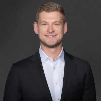 Professional portrait of a young man in a suit with a confident smile against a dark background.
