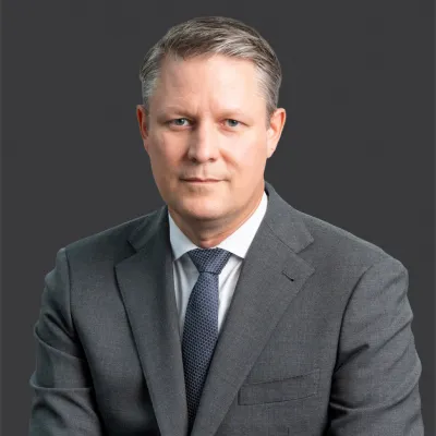 Portrait of a serious middle-aged man in a gray suit and blue tie against a dark gray background.