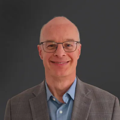 Smiling bald man wearing glasses, a gray blazer, and a blue shirt against a dark background.