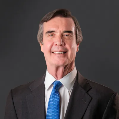 Professional portrait of a middle-aged man with gray hair in a dark suit and blue tie against a dark background.