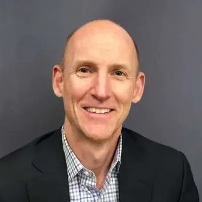 Smiling bald man in a checkered shirt and dark blazer against a plain gray background.