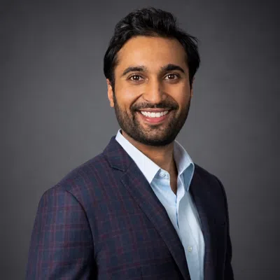 Smiling man in a blue plaid blazer and light blue shirt against a gray studio background