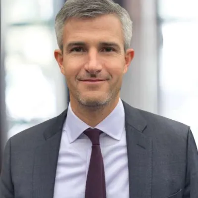 Confident middle-aged businessman with gray hair wearing a dark suit and maroon tie smiling in an office setting