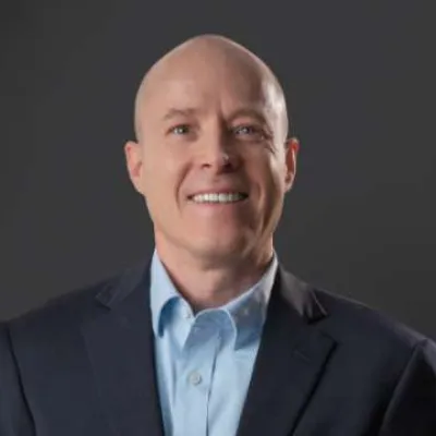 Smiling bald man in a dark suit jacket and light blue shirt against a dark background