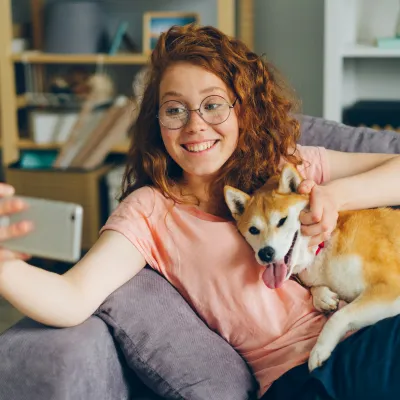 Smiling woman with glasses taking a selfie while holding a happy Shiba Inu dog on the couch at home