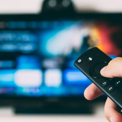Hand holding a black remote control pointed at a blurred smart TV screen with streaming app icons.