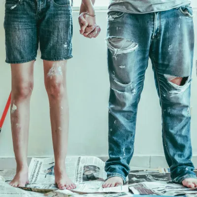Couple holding hands standing barefoot on newspapers with paint stains, holding paintbrushes in a room being painted
