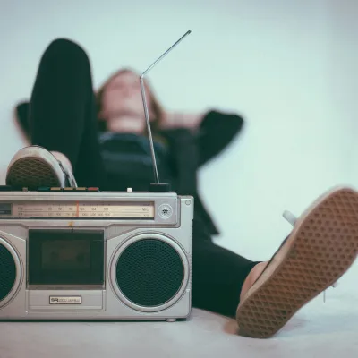 Vintage silver boombox with antenna in focus, person relaxing in background wearing black with sneakers visible.