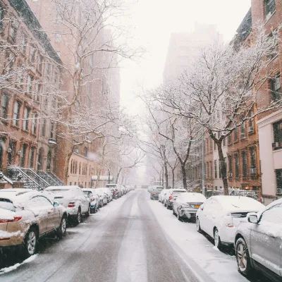 Snow-covered cars and bare trees line a city street with brownstone buildings in winter daylight.