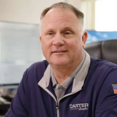 Man in a Carter Lumber jacket with American flag patch sitting in an office chair with a blurred office background.