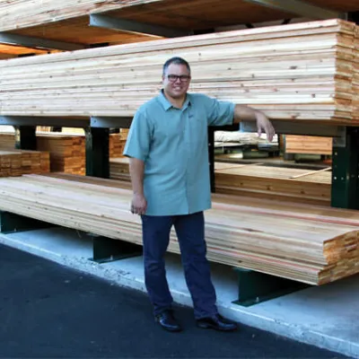 Man standing next to stacked lumber planks in an outdoor storage area of a lumber yard