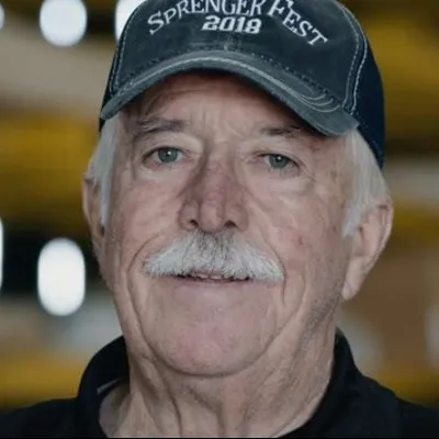 Smiling elderly man with a white mustache wearing a Sprenger Fest 2018 cap in a blurred industrial background