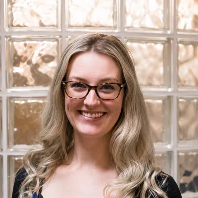 Smiling woman with blonde hair and glasses wearing a patterned top and black cardigan in front of glass blocks.