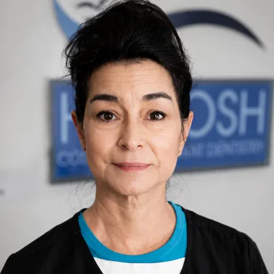 Professional middle-aged female dentist in black coat standing in front of dental clinic sign.