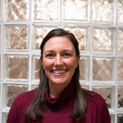 Smiling woman with long brown hair wearing a burgundy shirt in front of a glass block wall background