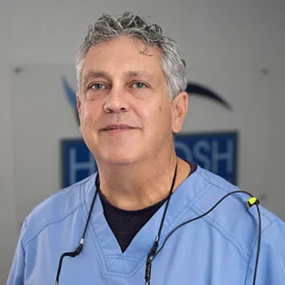 Middle-aged male dentist in blue scrubs smiling with dental equipment around his neck in a clinic setting.
