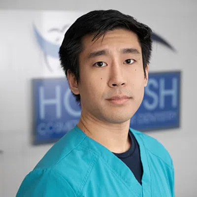 Young male healthcare professional in teal scrubs standing indoors with a blurred medical logo background
