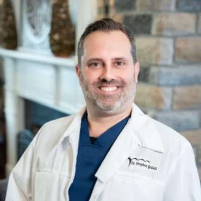 Smiling male doctor in white coat and navy scrubs standing in a room with stone wall and fireplace.