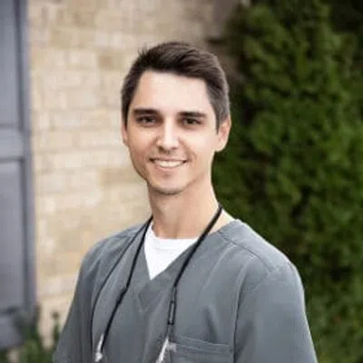 Smiling male healthcare professional wearing grey scrubs and a stethoscope outdoors with blurred greenery background.