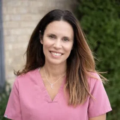 Smiling woman with long brown hair wearing pink medical scrubs outdoors near greenery and brick wall.