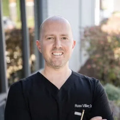 Smiling male doctor with arms crossed wearing black scrubs outdoors near a building with blurred background.