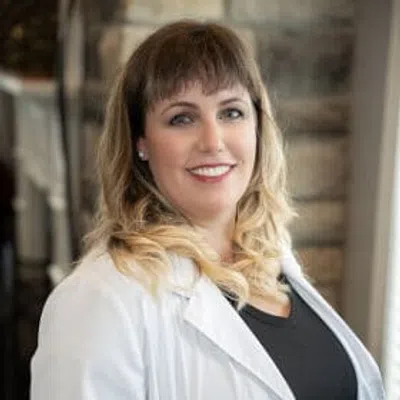 Smiling pregnant woman wearing white coat and black dress standing indoors near window with stone wall background