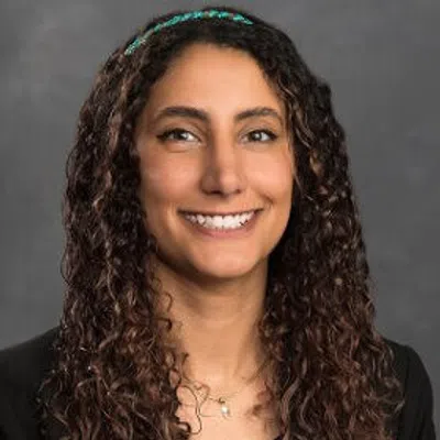 Smiling woman with long curly hair wearing a black blazer and polka dot blouse against gray background