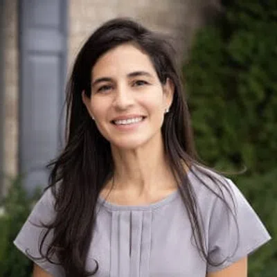 Smiling woman with long dark hair wearing a light gray blouse standing outdoors with greenery in the background