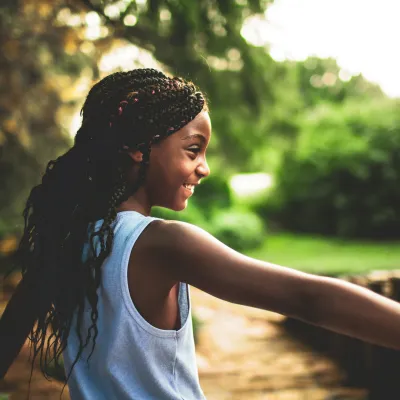 Smiling young girl with braided hair and white tank top enjoying nature walk on a sunny day.