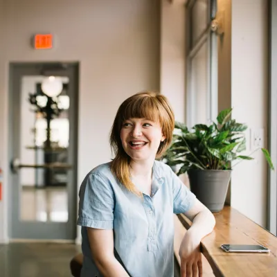 Smiling woman in a light blue shirt sitting by a window in a bright modern cafe with plants and natural light.