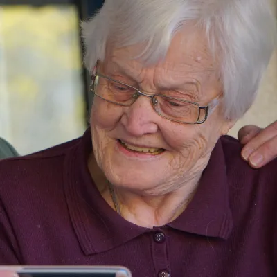 Elderly woman smiling while looking at a smartphone held by a masked caregiver with a comforting hand on her shoulder.