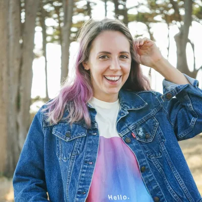 Smiling woman with purple hair wearing a denim jacket and colorful shirt saying Hello in an outdoor forest setting.
