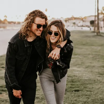 Young couple wearing sunglasses embracing and smiling while walking on grassy beachside path at sunset