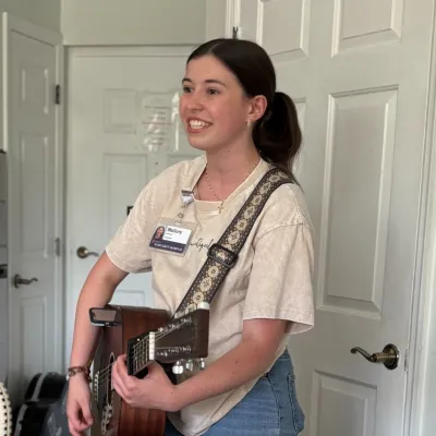 Young woman playing acoustic guitar indoors, smiling, next to wicker chairs and closed white doors.