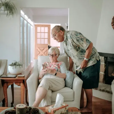 Elderly woman sitting on armchair using smartphone while man leans over to look at the screen in living room