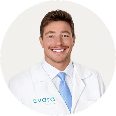 Smiling male doctor in white coat with blue tie and Evara Health logo on coat against white background.