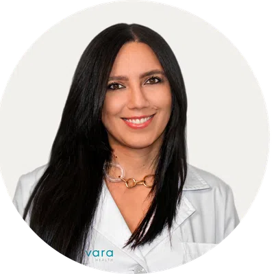 Portrait of a smiling female healthcare professional with long black hair wearing a white coat and gold necklace.