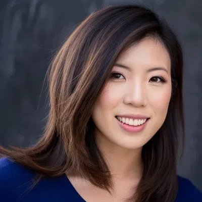 Smiling Asian woman with medium-length brown hair wearing a blue top against a dark blurred background.