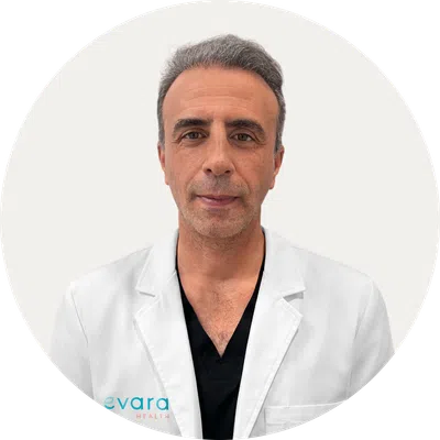 Middle-aged male doctor wearing a white coat and black scrubs with gray hair against a plain background.